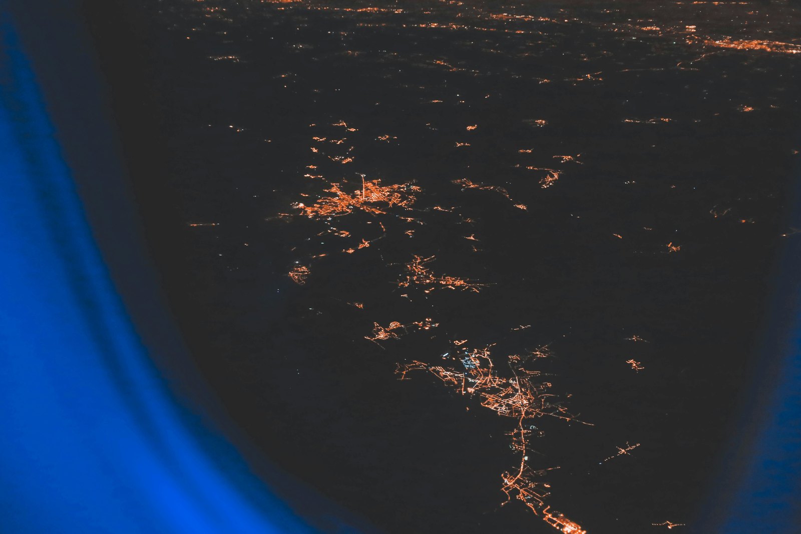 View through an aeroplane window at night, with scattered orange city lights spread across the dark ground below and the blue edge of the window frame in the foreground.