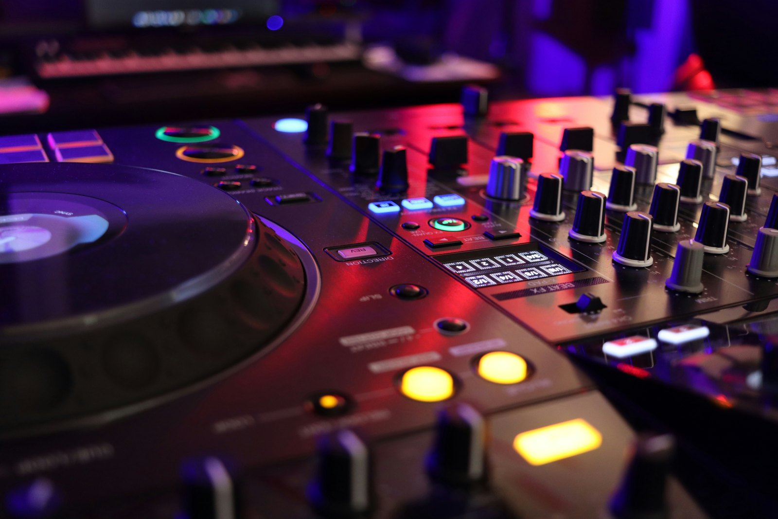 Close-up of a DJ mixer and the edge of a media player lit in deep red and purple, with glowing buttons and rows of knobs in a dark booth.