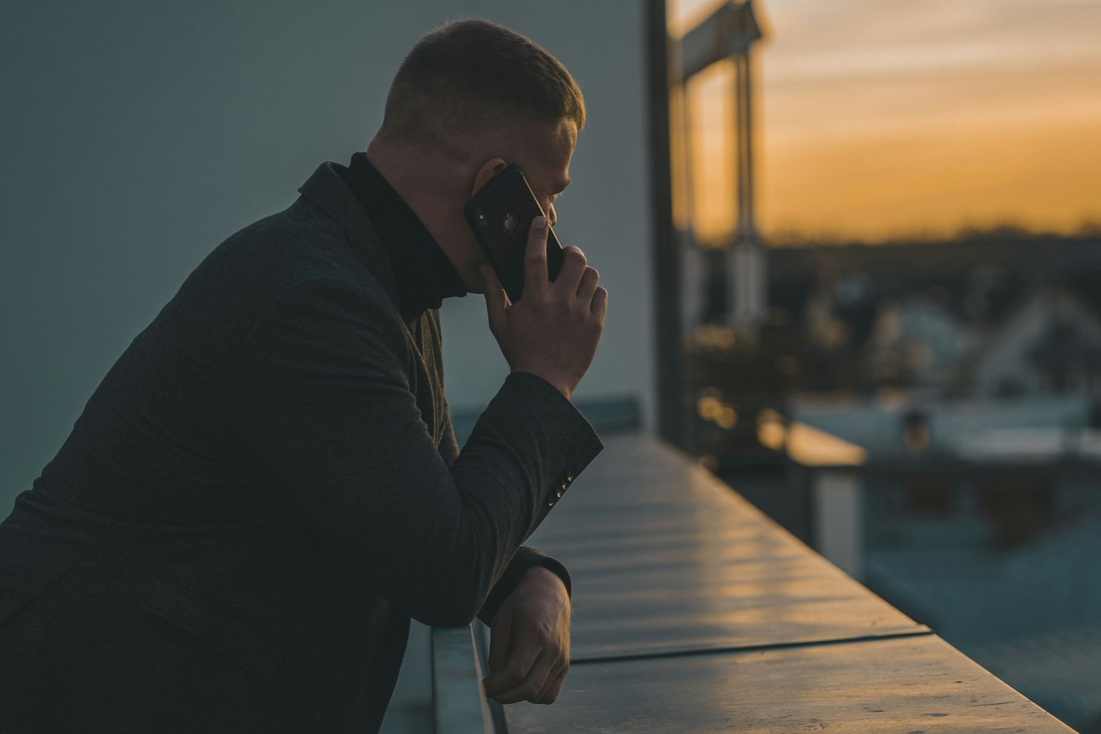 A man in a dark jacket leans on a rooftop ledge at dusk, talking on his phone, with blurred city rooftops and an orange sky behind him.
