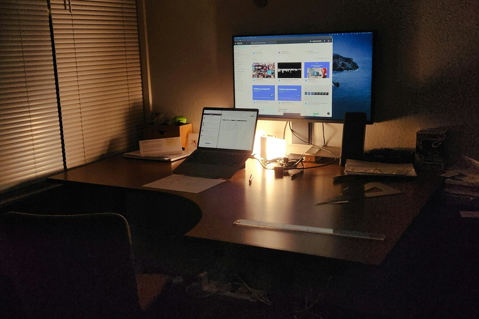 A dimly lit desk at night with a laptop and a monitor glowing under a desk lamp.