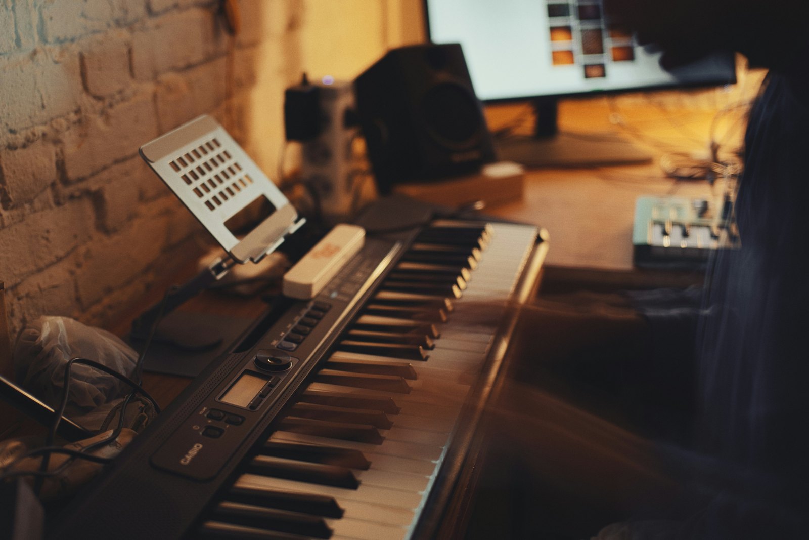 A person playing a digital keyboard in a dimly lit home studio, surrounded by synthesizers in warm low light.