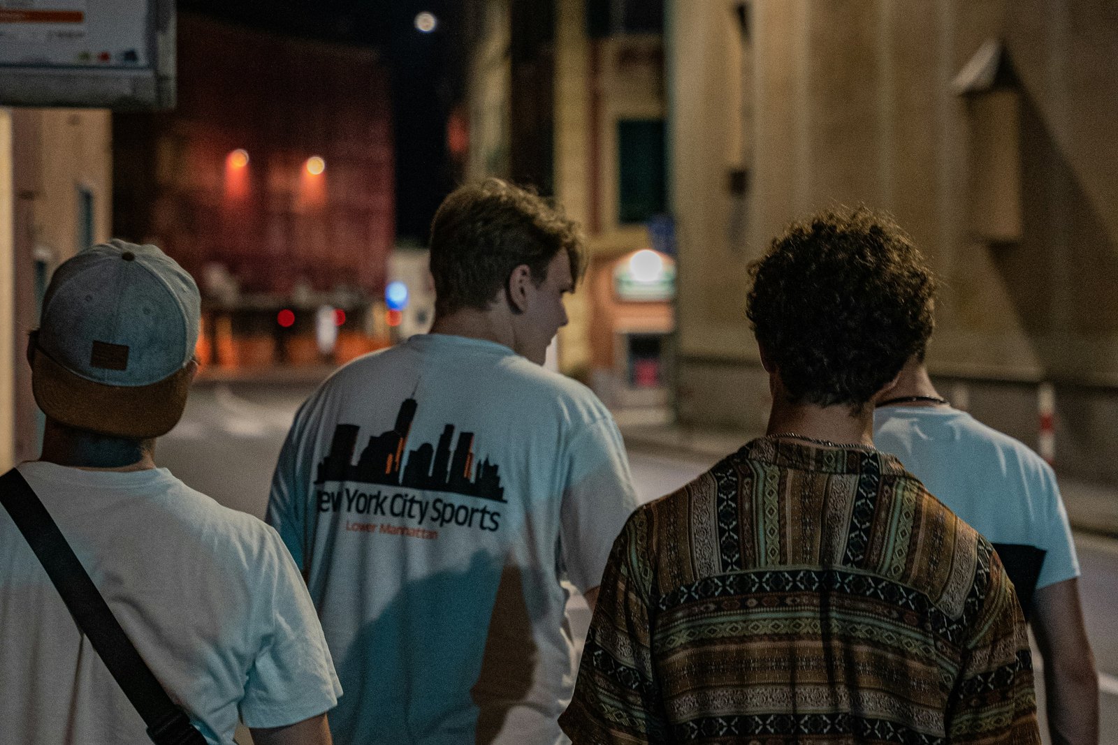 A group of people walking together down a city street at night, lit by warm streetlights and shopfronts.