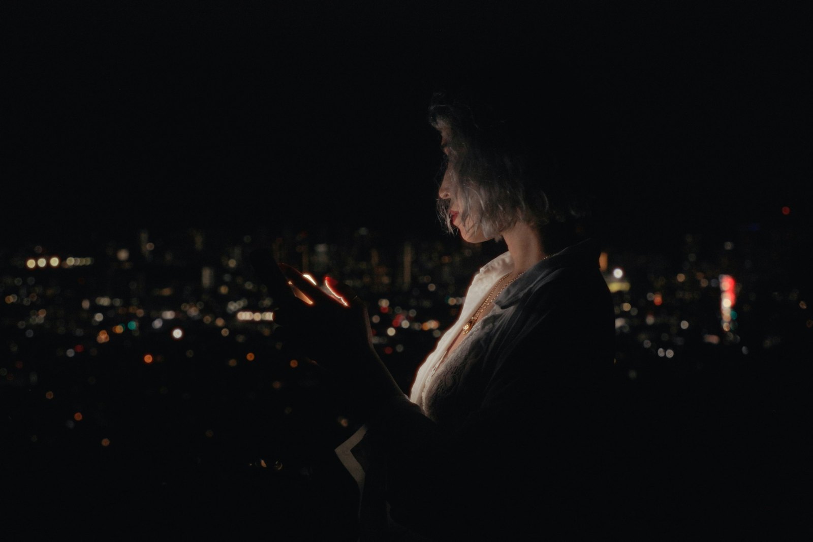 A woman standing in the dark at night, her face lit by her phone screen, with blurred city lights behind her.