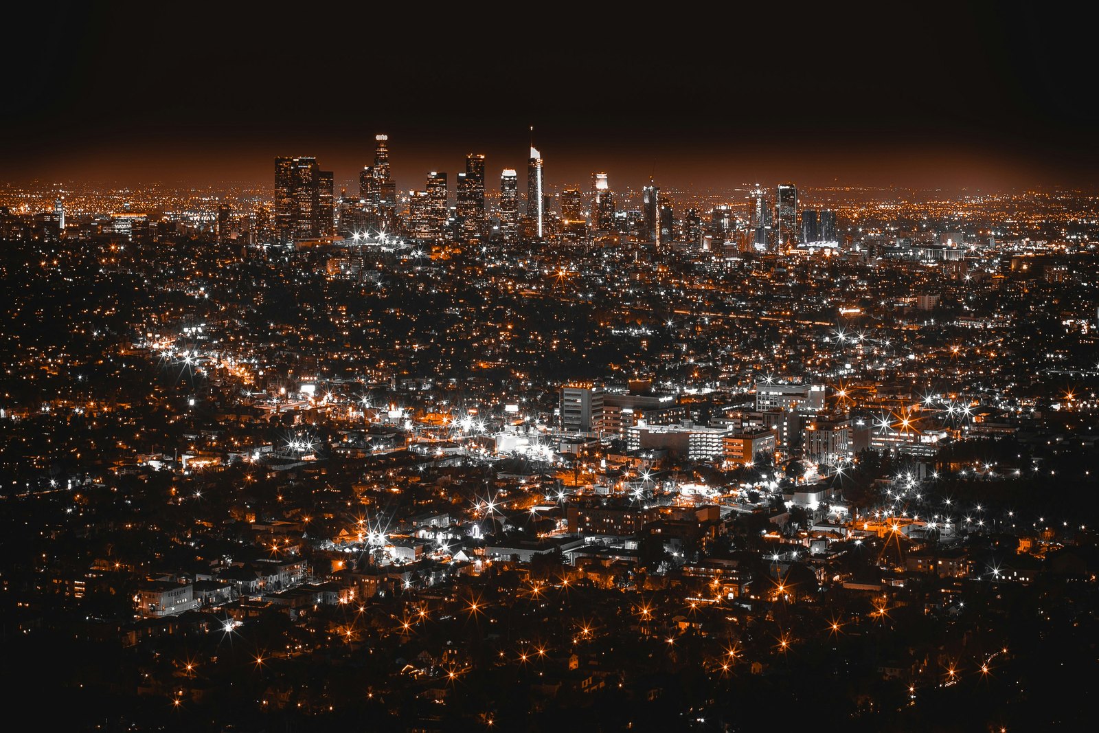 Night view over a sprawling city from a hillside, thousands of orange streetlights stretching toward a cluster of downtown towers on the horizon.