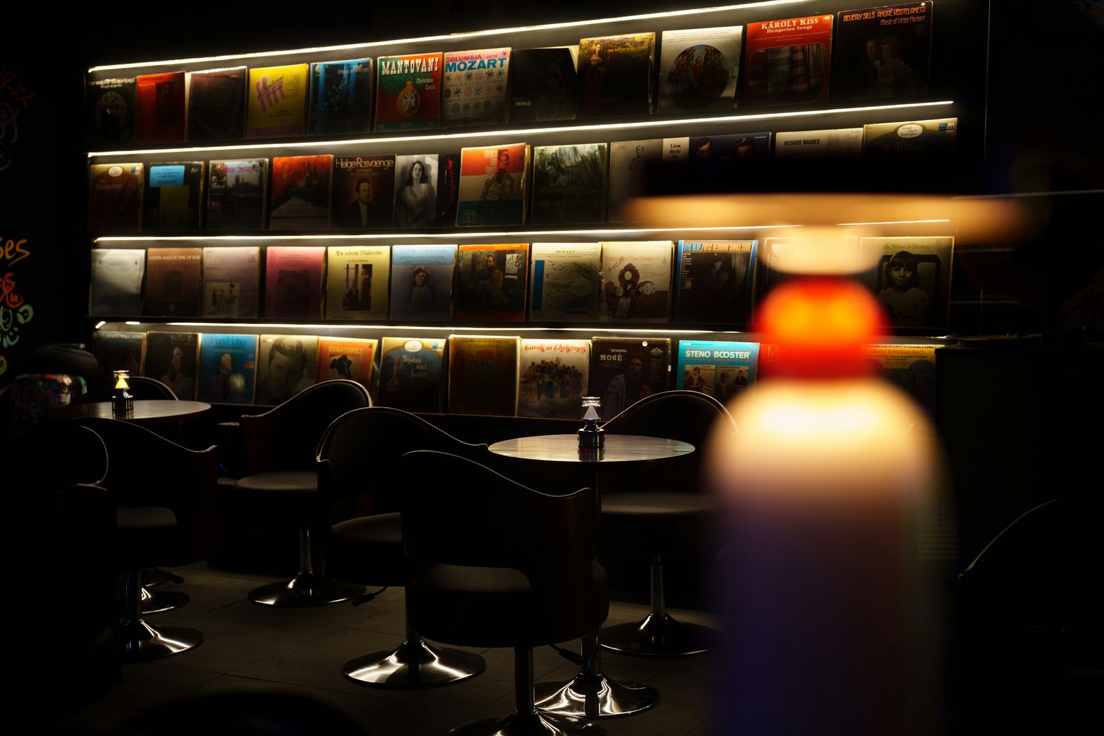 A dimly lit listening bar with a backlit wall of vinyl records, small round tables and a warm glowing lamp in the foreground.
