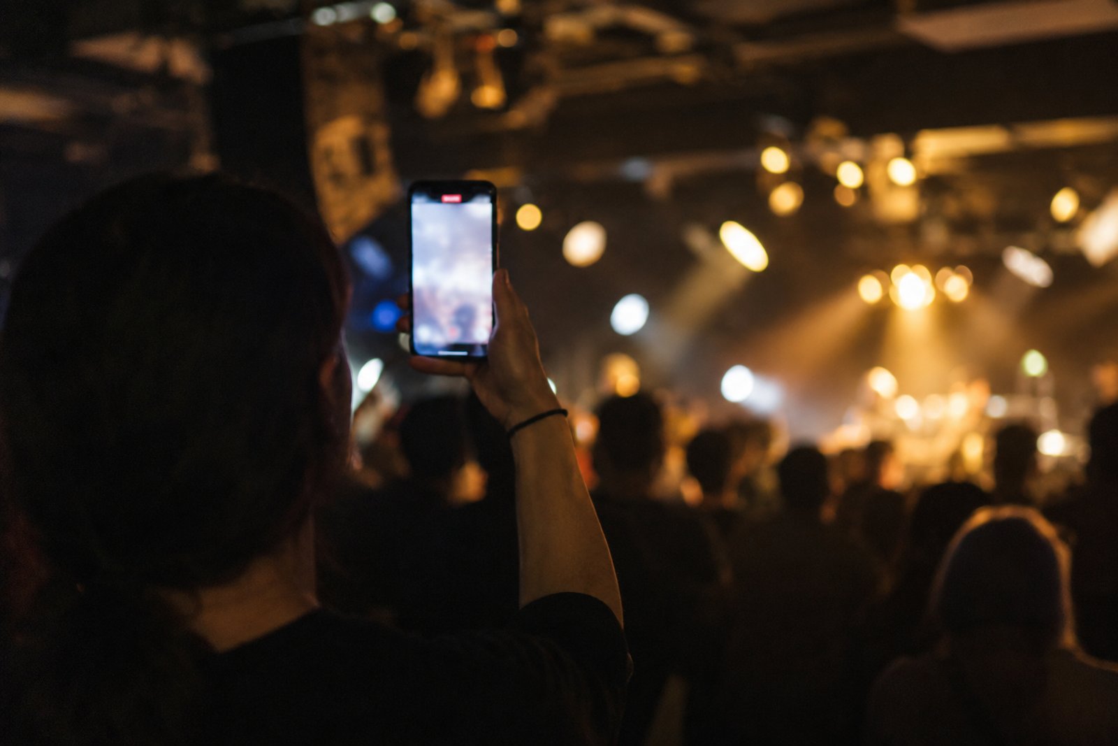 Fan in a dark club holding up a glowing phone toward the DJ booth