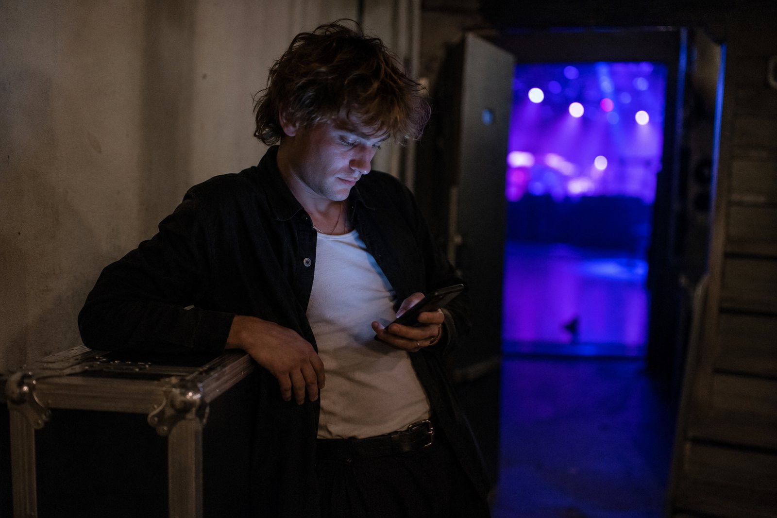 Musician backstage reading Instagram DMs on a phone, stage lights glowing behind
