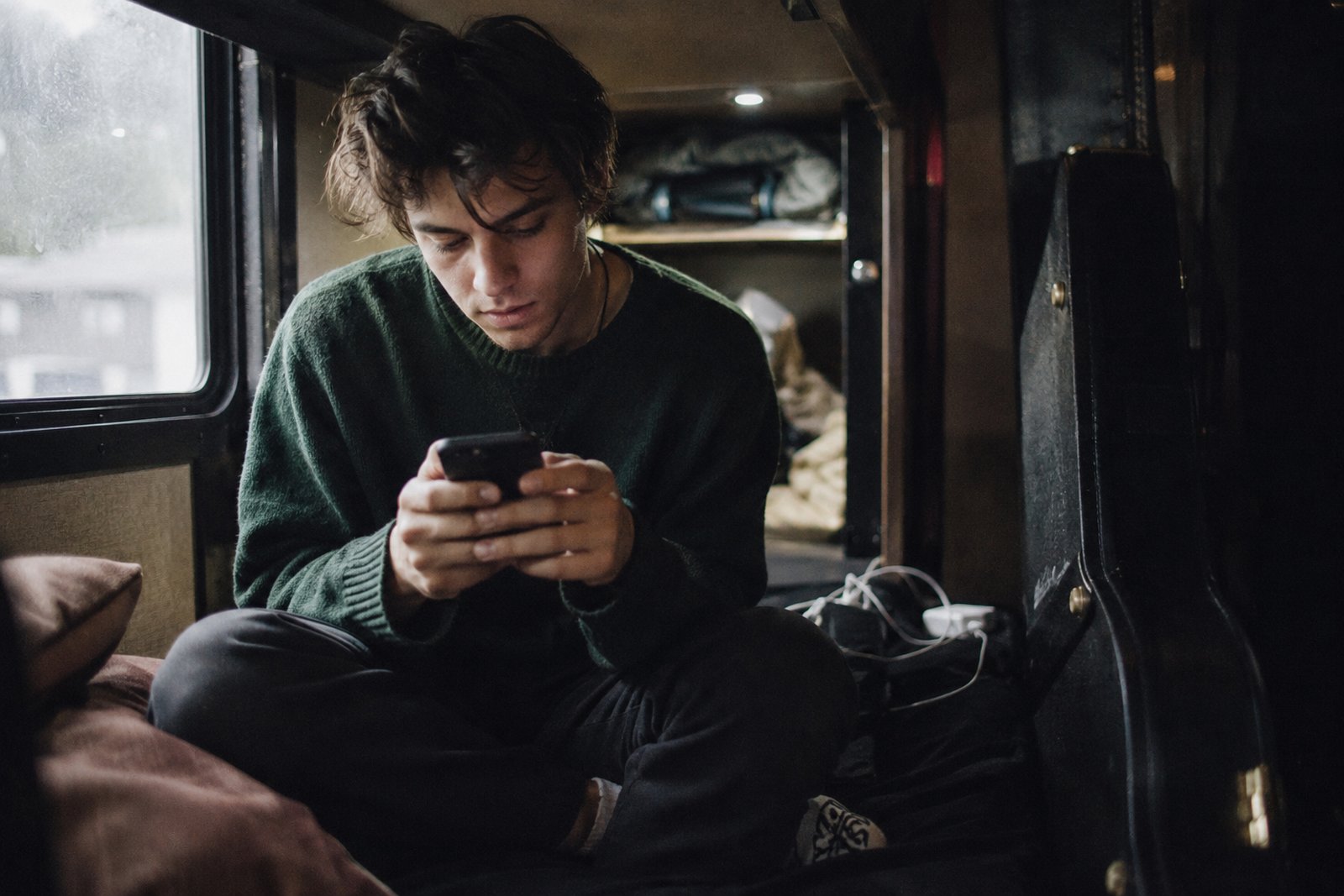 Musician sitting on a tour-bus bunk typing a post on a phone, soft window light